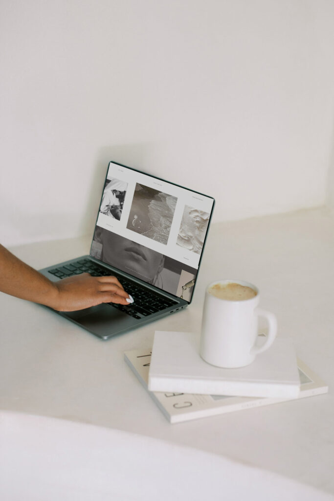 Best SEO for Business Coaches | High Tide Strategy Full Service Marketing Agency | A person with manicured nails uses a laptop displaying monochrome images. Nearby, a white coffee mug sits on stacked magazines, creating a calm, minimalist vibe.