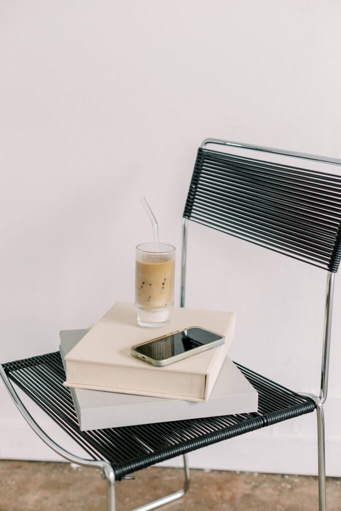 SEO Tips for Photographers | High Tide Strategy Full Service Marketing Agency | A modern chair with a black wire design holds stacked books, a smartphone, and an iced coffee in a glass with a straw, creating a minimalist, calm vibe.