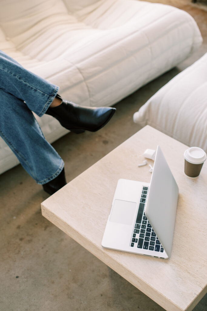 Coaching Website SEO | High Tide Strategy Full Service Marketing Agency | A laptop sits partially open on a light wooden table next to a to-go coffee cup. In the background, a person in jeans and black boots is seated on a white couch.