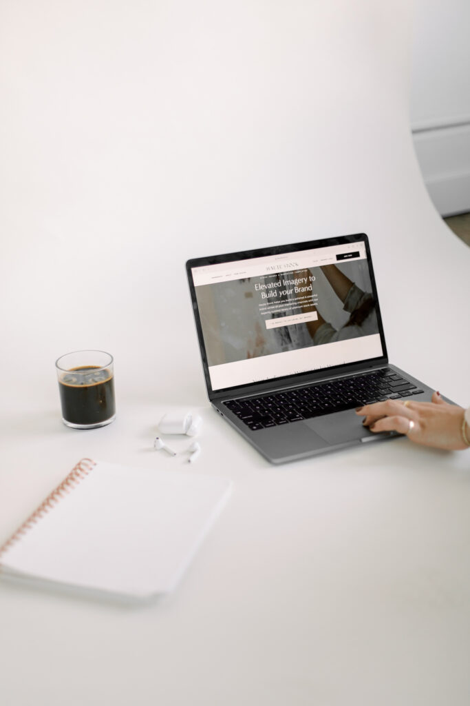Ecommerce Marketing Solutions | High Tide Strategy Full Service Marketing Agency | A hand types on a sleek laptop displaying a website about brand imagery. Nearby, there's a notebook, AirPods, and a glass of dark coffee on a white desk.