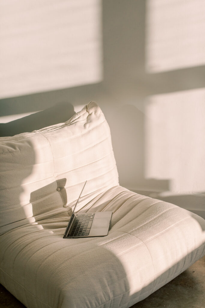 Showit Templates for Creatives | High Tide Strategy Full Service Marketing Agency | A laptop rests open on a cream-colored sofa. Soft sunlight streams through a window, casting gentle shadows on the sofa, creating a calm, serene ambiance.