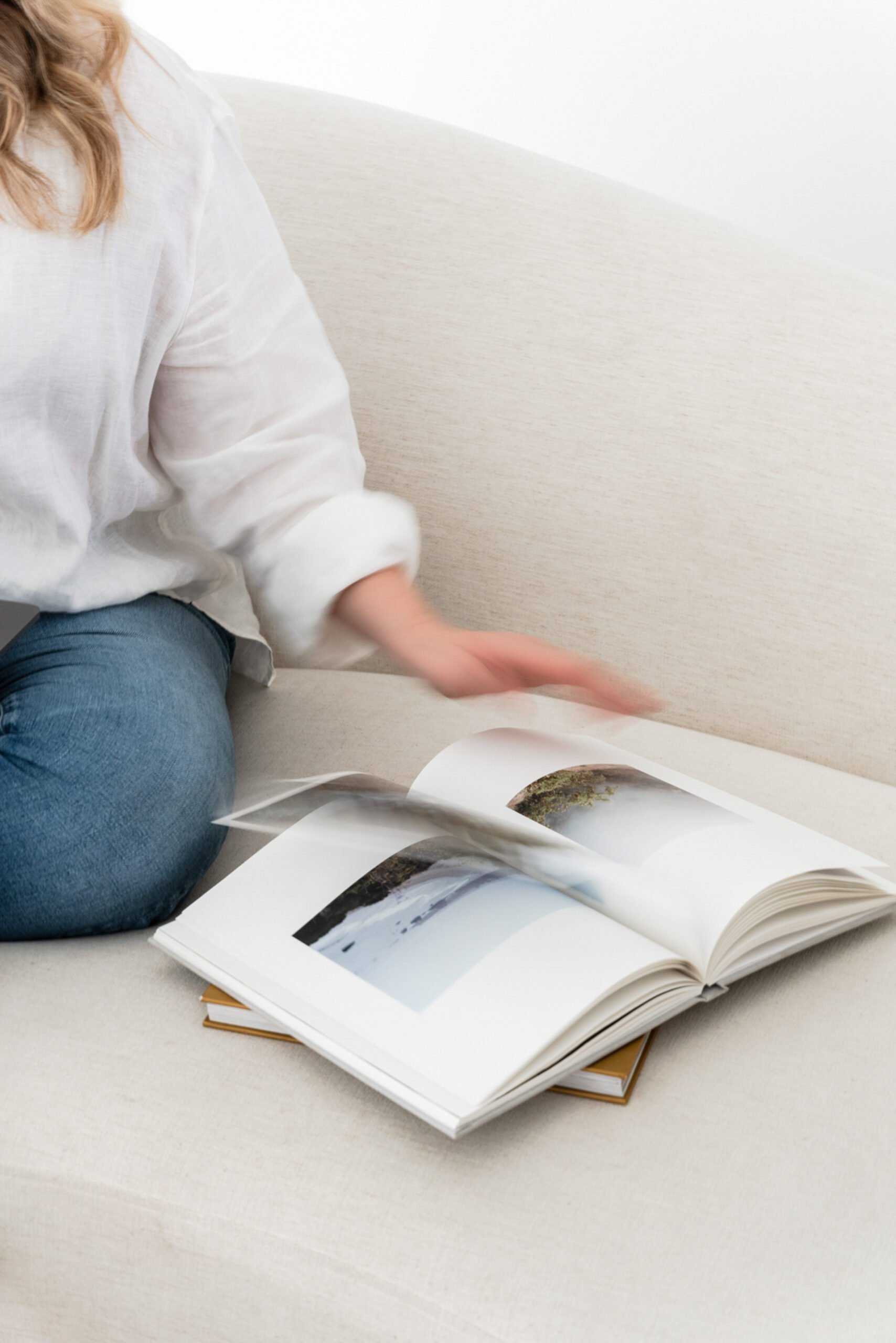 Heirloom Albums | High Tide Strategy Full Service Marketing Agency | A person in a white shirt and jeans sits on a beige couch, flipping through a photo book with landscape images. The scene feels calm and leisurely.