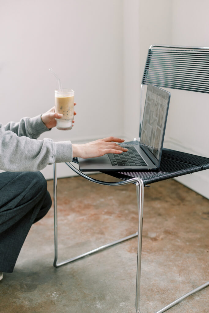 Showit Website Designs | High Tide Strategy Full Service Marketing Agency | Person in a gray sweater holds an iced coffee while using a laptop on a modern chair. The setting is minimalistic and the mood is focused and serene.