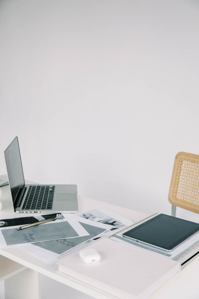 Best SEO for Photographers | High Tide Strategy Full Service Marketing Agency | A minimalist workspace features an open laptop, scattered papers, a pen, and an AirPod case on a white desk. A wicker chair creates a calm, organized vibe.