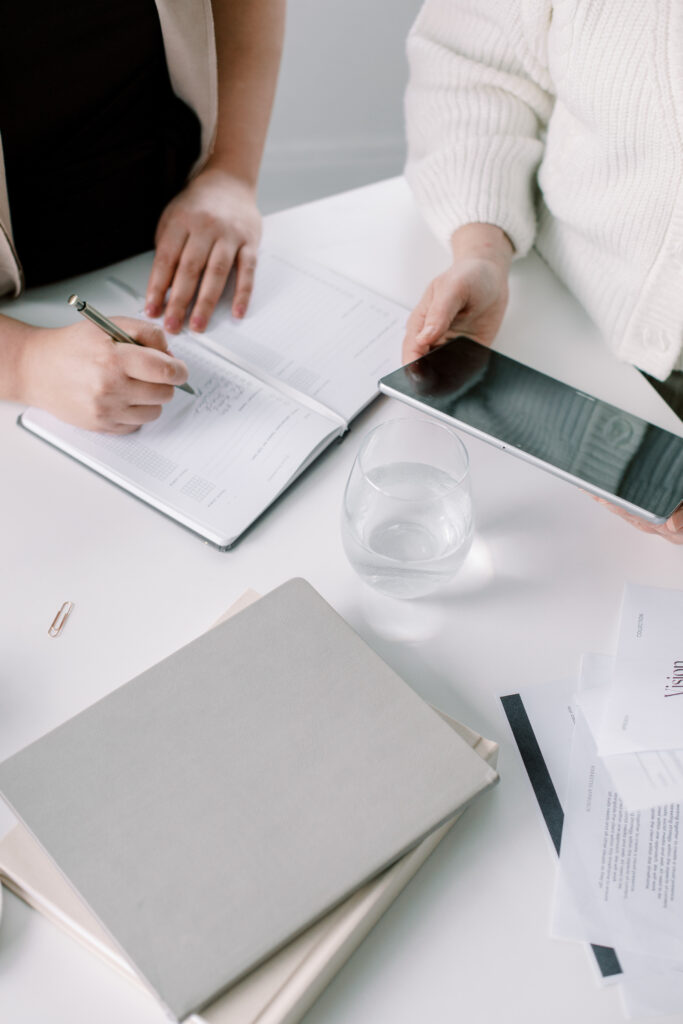 SEO Course for Photographers | High Tide Strategy Full Service Marketing Agency | Two people collaborate at a white table with a notebook, tablet, and glass of water. One writes notes, evoking a focused, productive atmosphere.