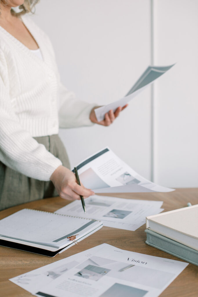 SEO Tips for Photographers | High Tide Strategy Full Service Marketing Agency | A person in a white sweater organizes paperwork on a wooden table, surrounded by open books and documents. The scene conveys a busy, productive atmosphere.