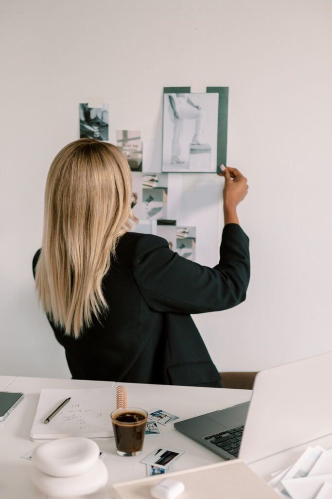SEO Services for Coaches | High Tide Strategy Full Service Marketing Agency | A woman with long blonde hair, wearing a black blazer, arranges photos on a white wall. Her workspace has a laptop, notebook, and coffee cup, suggesting creativity.