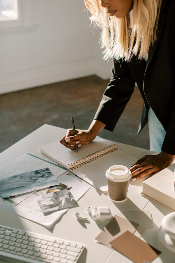 Best SEO for Photographers | High Tide Strategy Full Service Marketing Agency | Woman with long blonde hair writing in a spiral notebook at a sunlit desk with a coffee cup, photos, stack of papers, and pen scattered around.