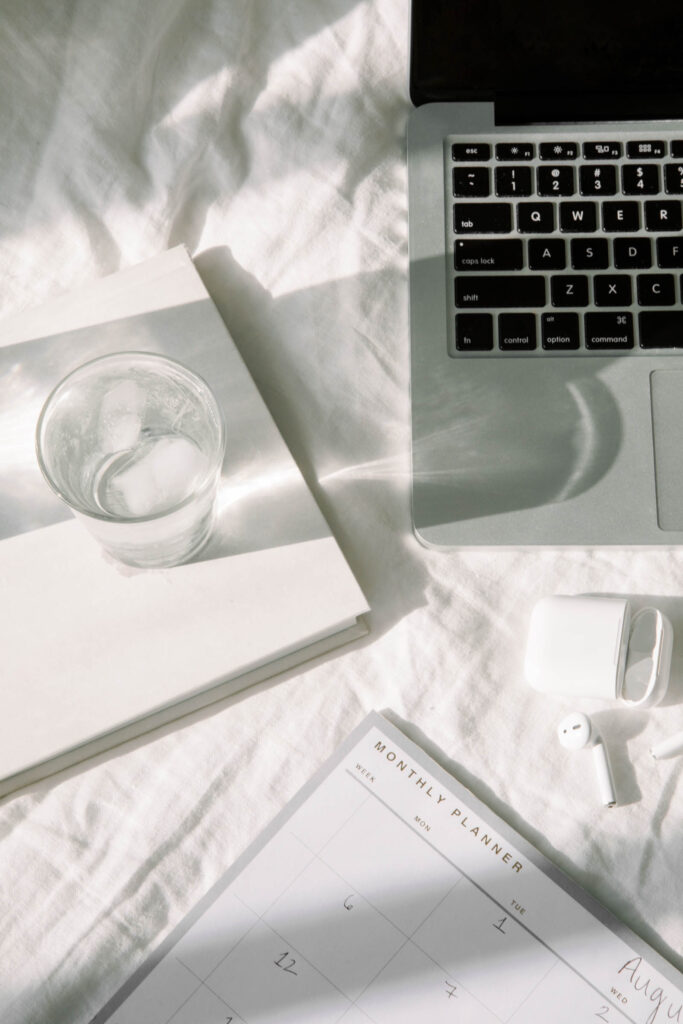 SEO Expert for Photographers | High Tide Strategy Full Service Marketing Agency | A minimalist workspace features a laptop on light bedding, a clear glass with ice on a book, and an open planner with earbuds. The scene is calm and organized.