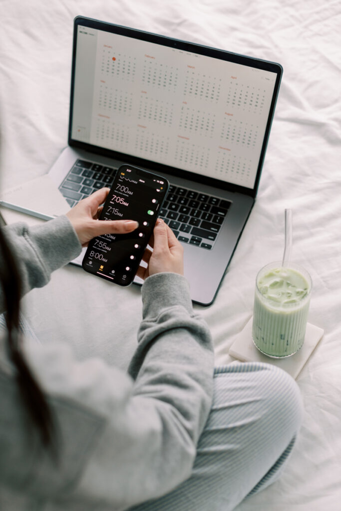 SEO Services for Photographers | High Tide Strategy Full Service Marketing Agency | A person in gray pajamas checks alarms on a smartphone while sitting with a laptop displaying a calendar. A green smoothie is nearby on a white bed.