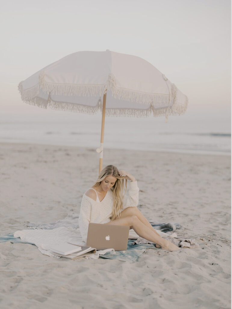 Best Local SEO Keywords for Business Coaches | High Tide Strategy Full Service Marketing Agency | Mandy in a white sweater sits on a blanket under a fringed beach umbrella, using a laptop. The serene beach setting conveys a relaxed atmosphere.