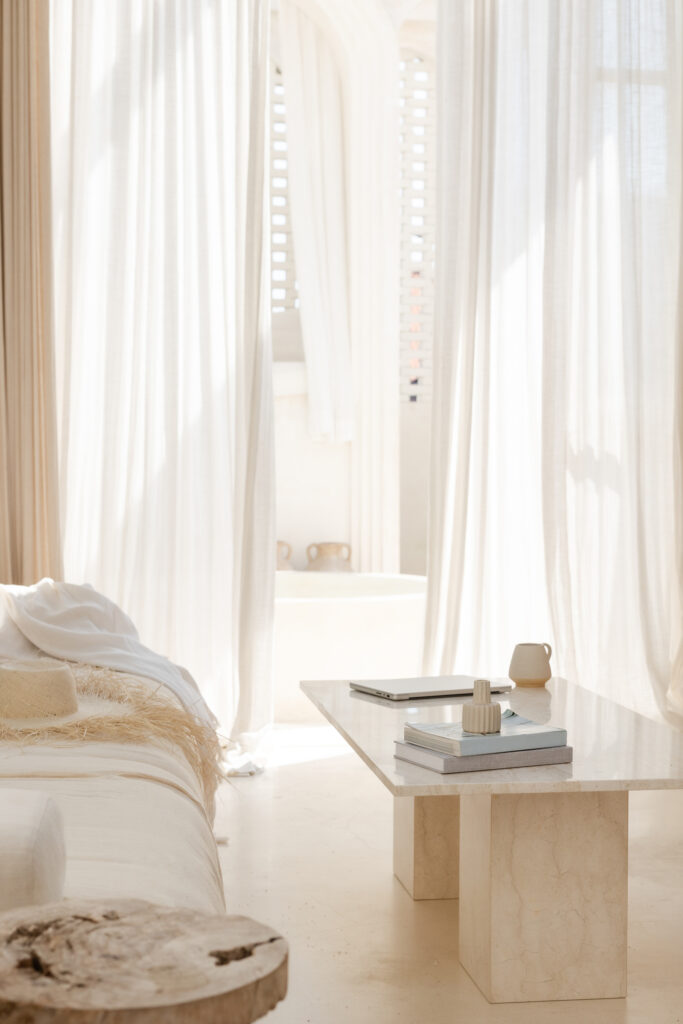 Interior Designer Marketing Plan | High Tide Strategy Full Service Marketing Agency | Serene room with soft beige tones. A cozy sofa with a throw faces a stone coffee table with books, a laptop, and a mug. Sunlit curtains add warmth.