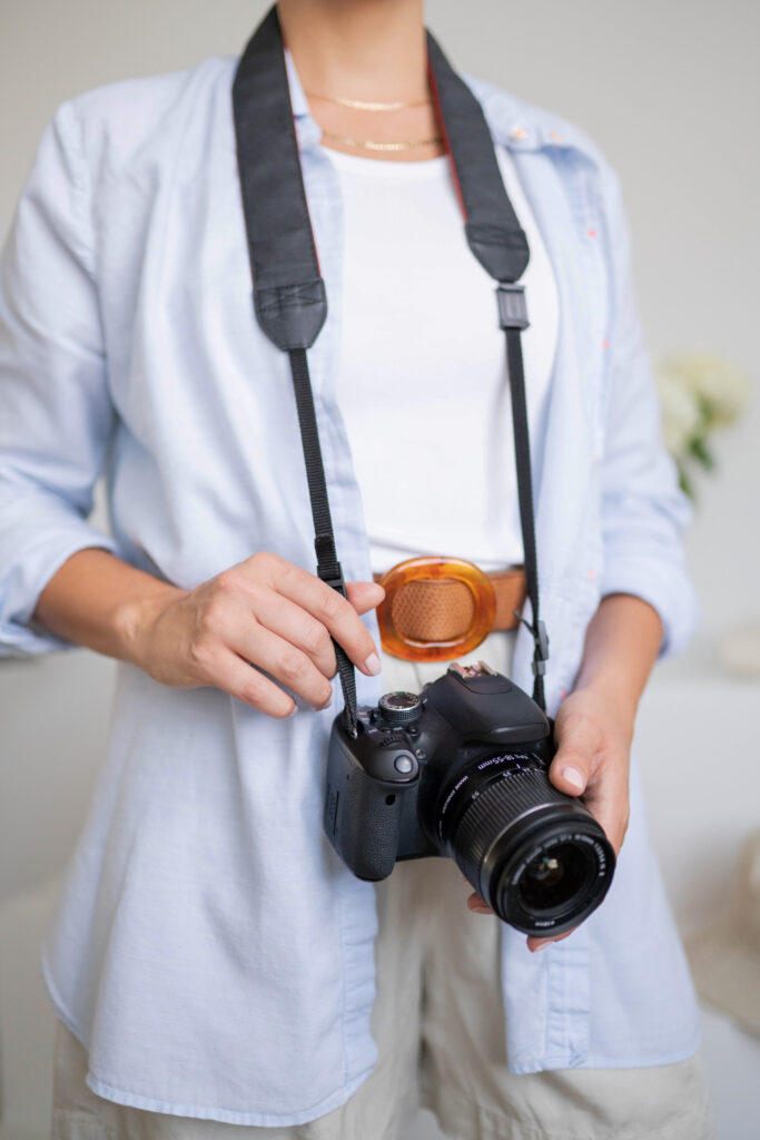 SEO Services for Photographers | High Tide Strategy Full Service Marketing Agency | Person in light blue shirt and white top holds a DSLR camera with a strap. A brown belt adds contrast. Soft focus background suggests a calm ambiance.