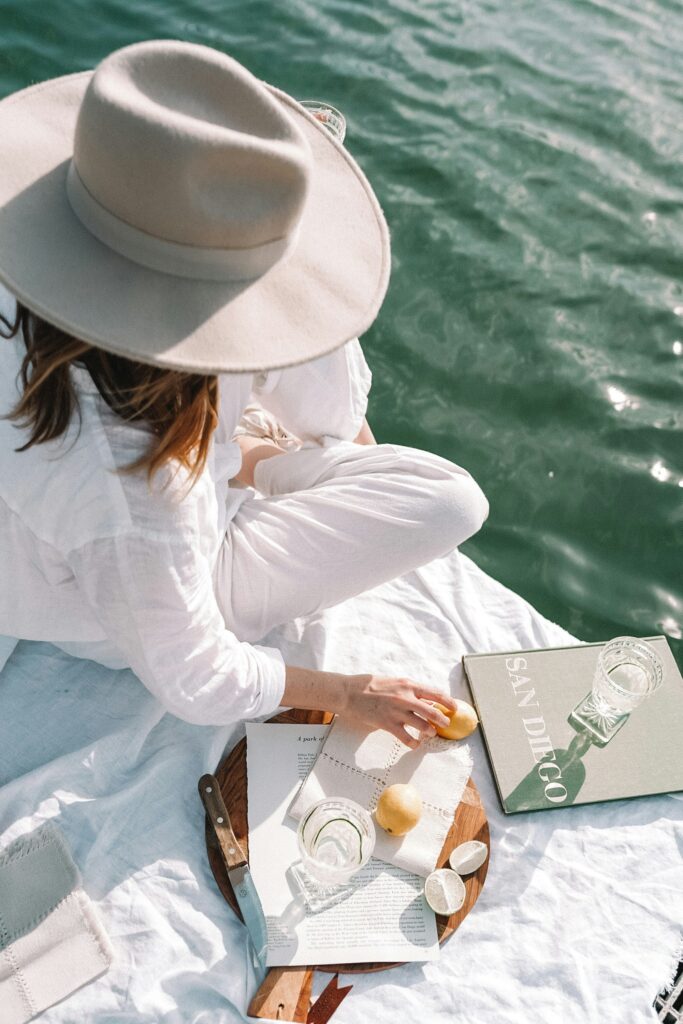 SEO Services for Wedding Photographers | High Tide Strategy Full Service Marketing Agency | A woman in a sun hat sits on a white blanket by the water, with lemons, a knife, and a book titled "San Diego" beside her. The scene is serene and leisurely.