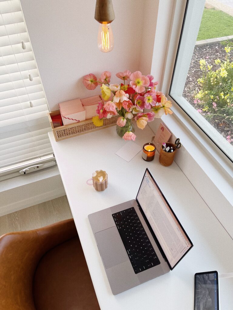 Florist Marketing Strategy | High Tide Strategy Full Service Marketing Agency | A cozy workspace with a laptop on a white desk by a window. A bouquet of pink and yellow flowers, a lit candle, and a mug add warmth and charm.