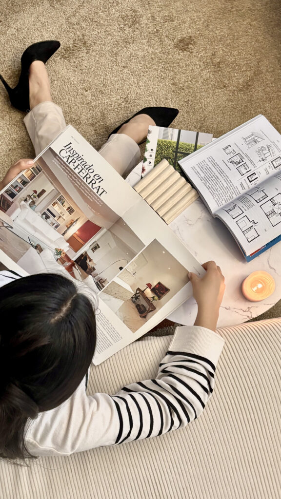 Interior Design Marketing Plan | High Tide Strategy Full Service Marketing Agency | Person sitting on carpet reading a magazine with interior design images. Nearby are open floor plans, fabric swatches, and a lit candle. Cozy and focused atmosphere.