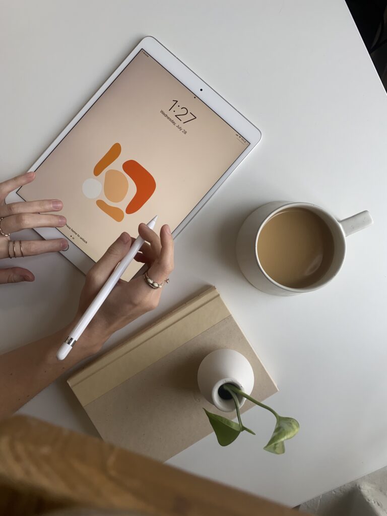 Content Marketing for Creatives | High Tide Strategy Full Service Marketing Agency | Hands holding a tablet with abstract art on the screen at 1:27 PM. Surrounding items include a coffee cup, a closed book, and a small vase with a green sprout.