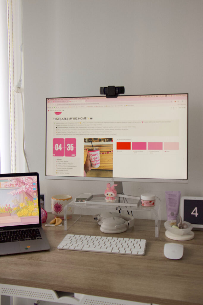 Online Marketing for Creative Entrepreneurs | High Tide Strategy Full Service Marketing Agency | A tidy desk setup with a large monitor displaying a website with pink accents. A laptop shows a floral background. Accessories include a webcam, keyboard, mouse, and cute figurines, creating a cozy, organized feel.