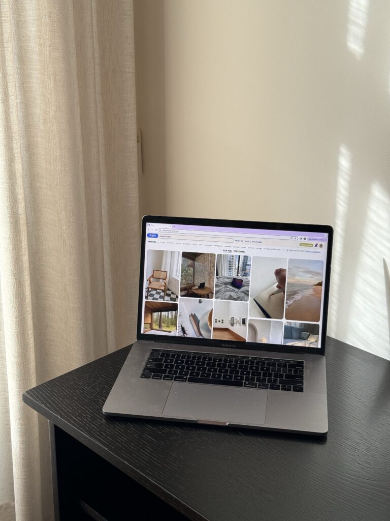 SEO for Shopify Stores | High Tide Strategy Full Service Marketing Agency | A laptop on a dark wooden table displays a Pinterest webpage featuring interior design images. Sunlight streams through a nearby curtain, creating a warm atmosphere.