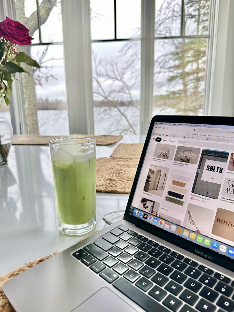 Online Boutique Marketing | High Tide Strategy Full Service Marketing Agency | A laptop on a table shows a Pinterest page. Beside it is a glass of iced green matcha. Large windows reveal a serene, overcast lake view.