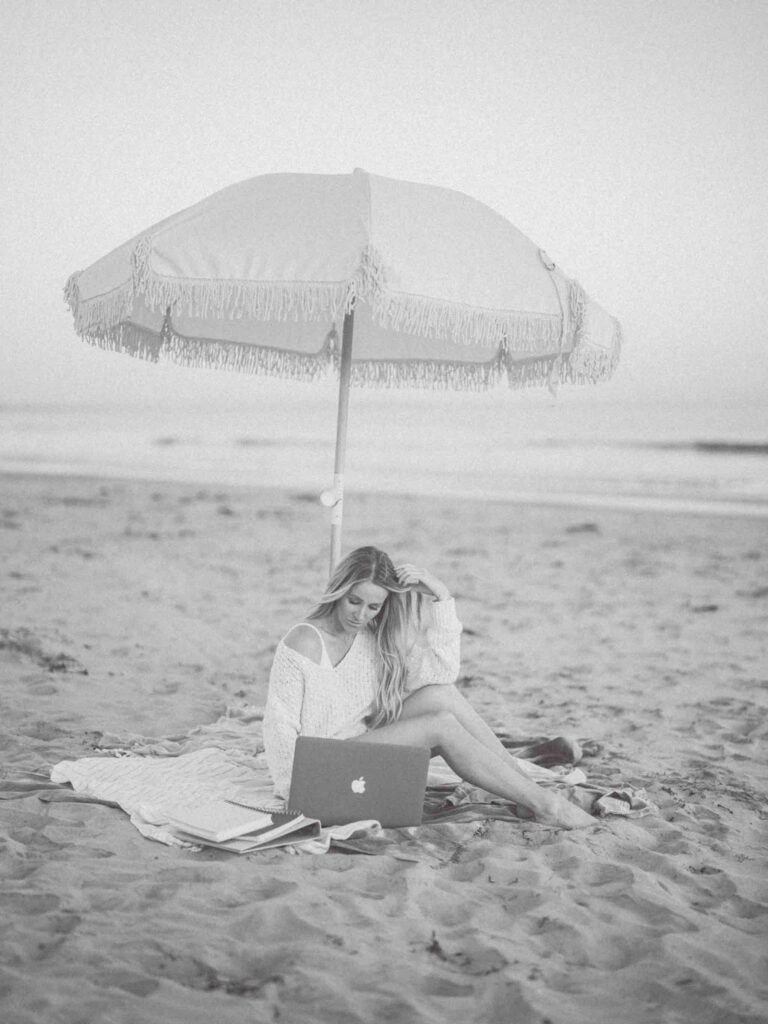 A woman from High Tide Strategy sits on the beach with her laptop open under a white umbrella