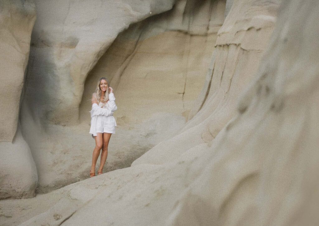 Mandy from High Tide Strategy stands in front of a big wall of sand while wearing white shorts and a white long sleeve shirt