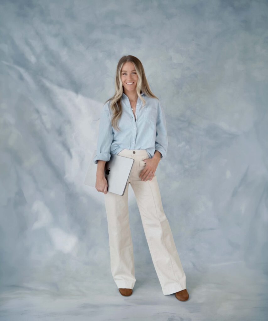 Mandy Ford | SEO Expert For Photographers, stands behind a blue backdrop with a laptop in hand.