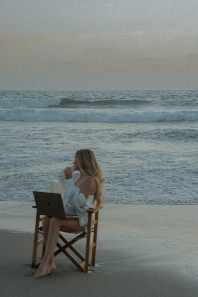 Mandy Ford of High Tide Strategy sits in a chair with a laptop on her lap on the beach in front of the ocean