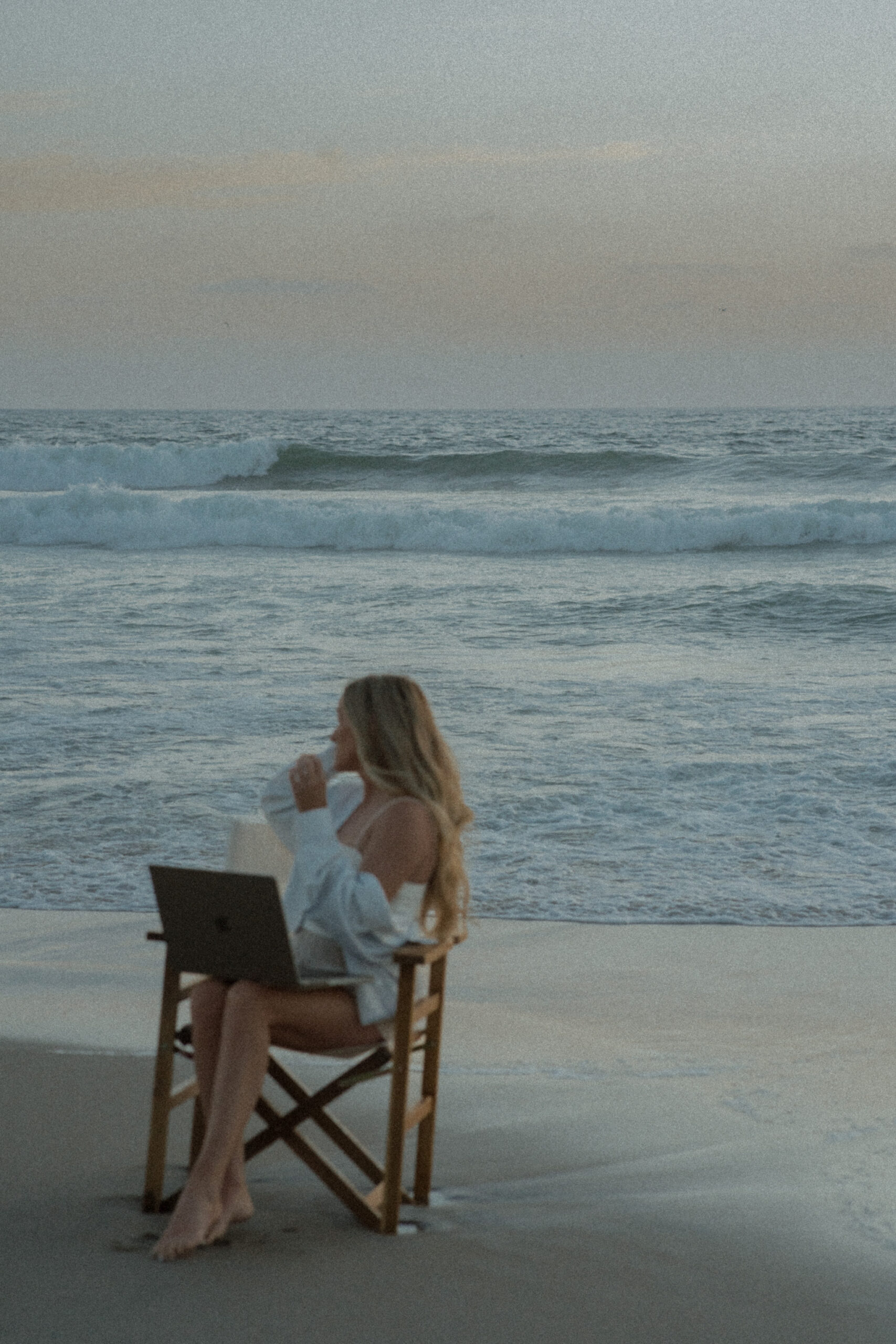 Mandy Ford of High Tide Strategy sits in a chair with a laptop on her lap on the beach in front of the ocean