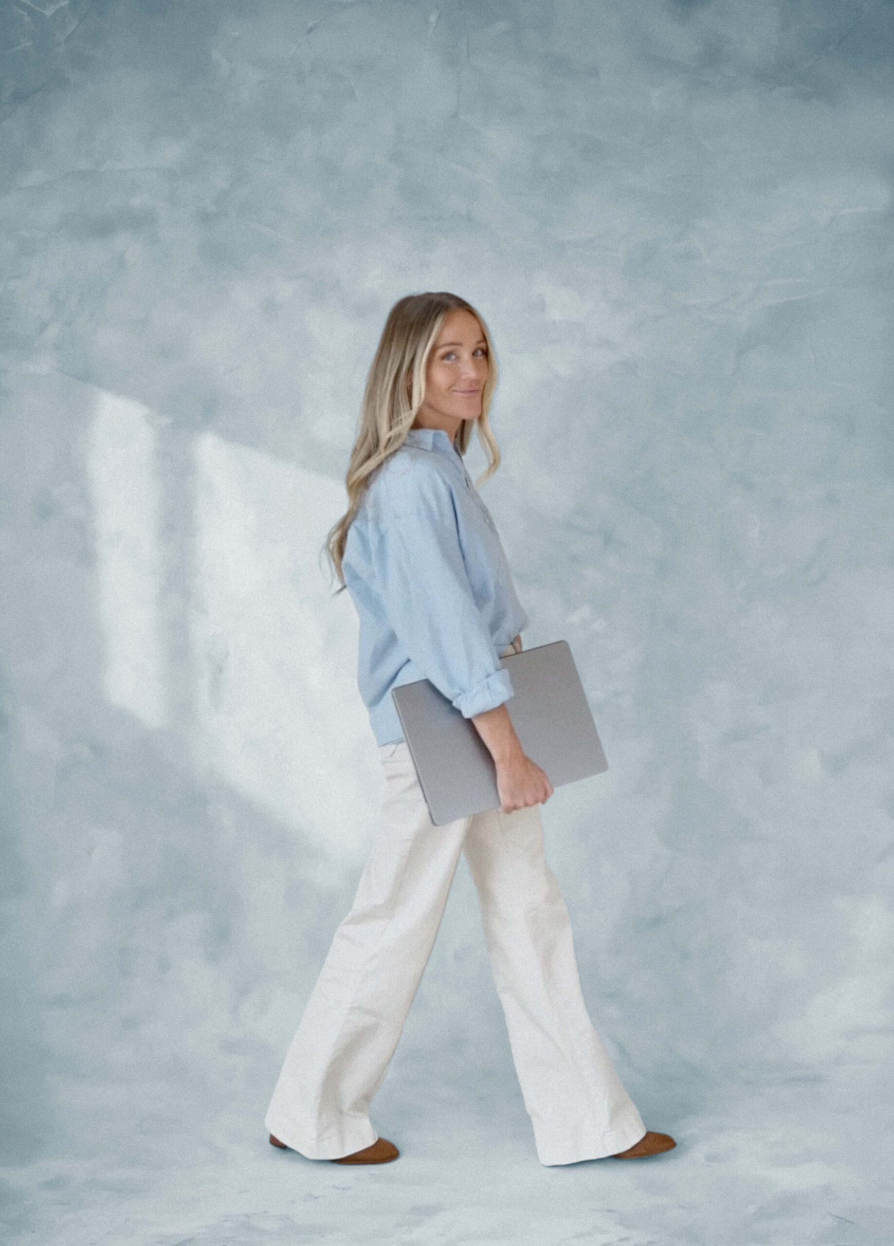 Mandy from High Tide Strategy stands in front of a textured background with a laptop in her hand wearing a blue long sleeve shirt